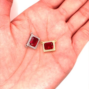 May include: Two rectangular gemstone pendants. One is silver-toned with a red center stone and a border of small clear stones. The other is gold-toned with a red center stone and a border of small clear stones. Both have small loops for attaching to a chain.