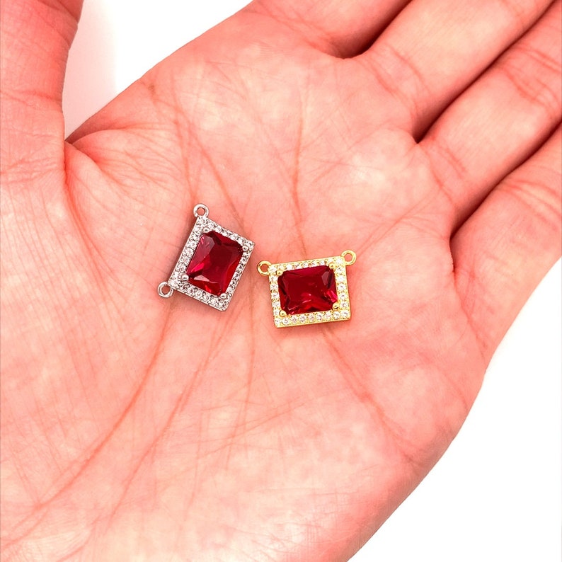 May include: Two rectangular gemstone pendants. One is silver-toned with a red center stone and a border of small clear stones. The other is gold-toned with a red center stone and a border of small clear stones. Both have small loops for attaching to a chain.