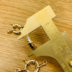 K&ouml;nnte beinhalten: Goldfarbene Schmuckverschl&uuml;sse mit Federringverschluss und ein Messwerkzeug. Die Verschl&uuml;sse haben ein kreisf&ouml;rmiges Design mit kleinen Verbindungs&ouml;sen. Das Messwerkzeug ist goldfarben mit schwarzen Markierungen. Der Hintergrund ist eine hellbraune Holzoberfl&auml;che.