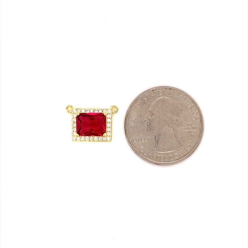 May include: A gold-colored pendant with a rectangular, deep red gemstone at its center, surrounded by smaller, clear stones. The pendant is shown next to a U.S. quarter for size comparison. The pendant has two small loops for attaching to a chain.
