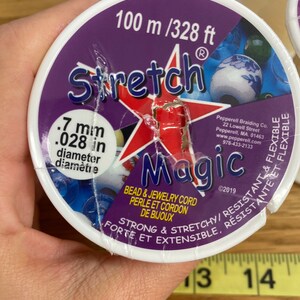 Puede incluir: Un carrete de cord&oacute;n Stretch Magic para cuentas y joyer&iacute;a. La etiqueta es morada con una estrella roja y texto blanco, incluyendo "100 m" y "0,7 mm de di&aacute;metro". El cord&oacute;n es fuerte, el&aacute;stico, resistente y flexible.