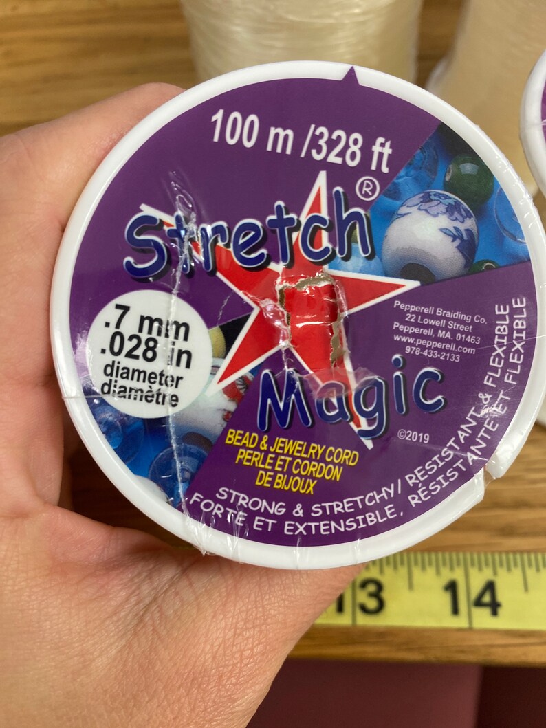 Puede incluir: Un carrete de cord&oacute;n Stretch Magic para cuentas y joyer&iacute;a. La etiqueta es morada con una estrella roja y texto blanco, incluyendo "100 m" y "0,7 mm de di&aacute;metro". El cord&oacute;n es fuerte, el&aacute;stico, resistente y flexible.