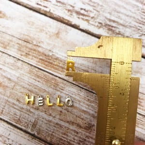 May include: Gold and silver letter charms spelling out the word "HELLO" with a gold letter "R" measured with a metal ruler.
