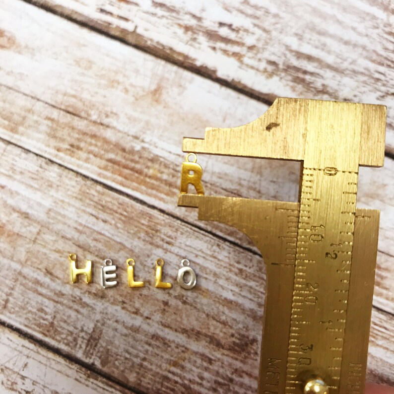 May include: Gold and silver letter charms spelling out the word "HELLO" with a gold letter "R" measured with a metal ruler.
