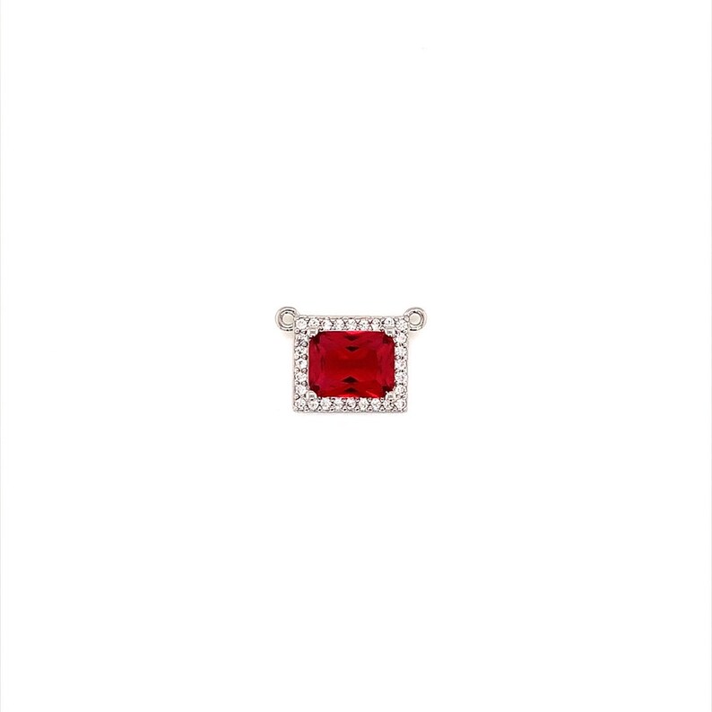 May include: A rectangular pendant featuring a large, vibrant red gemstone, surrounded by a halo of smaller, clear stones. The pendant is set in a silver-toned metal with small loops at the top for a chain.