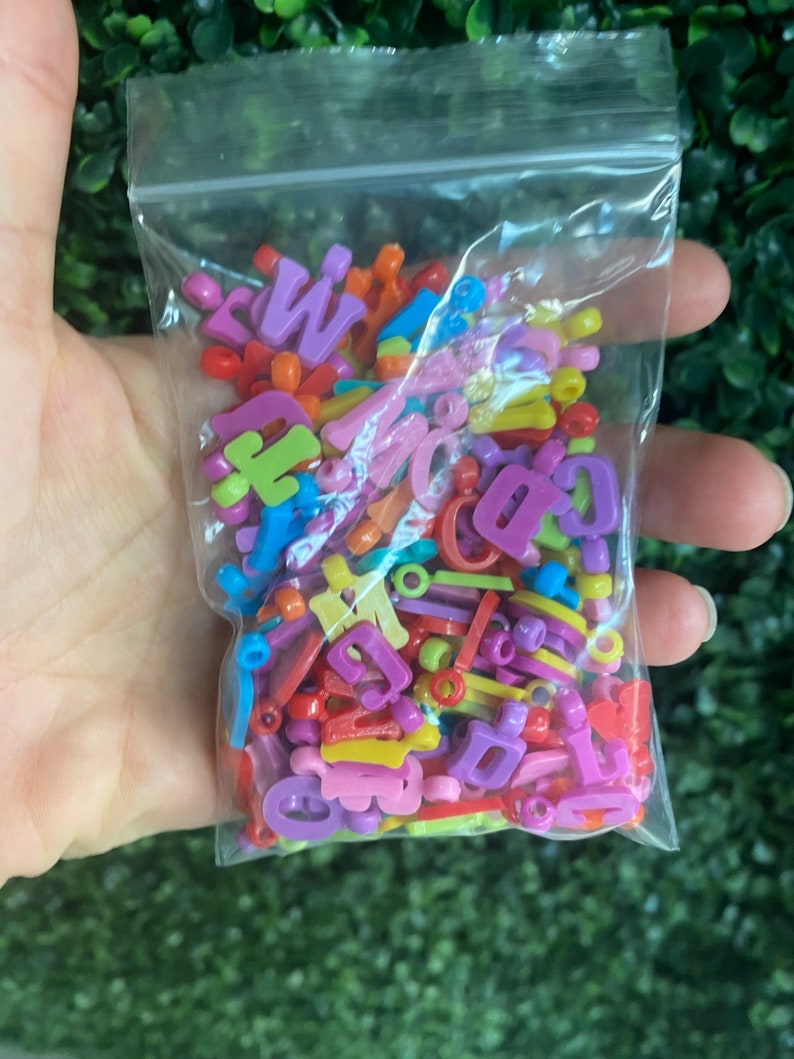 May include: A plastic bag filled with colorful letter beads in various shades of pink, purple, yellow, orange, green, and blue.
