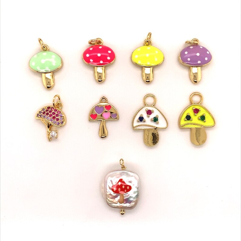 May include: A collection of gold-toned mushroom charms with colorful enamel caps. The charms feature various designs, including polka dots, hearts, and gemstones. One charm is a square pearl with a red mushroom design.