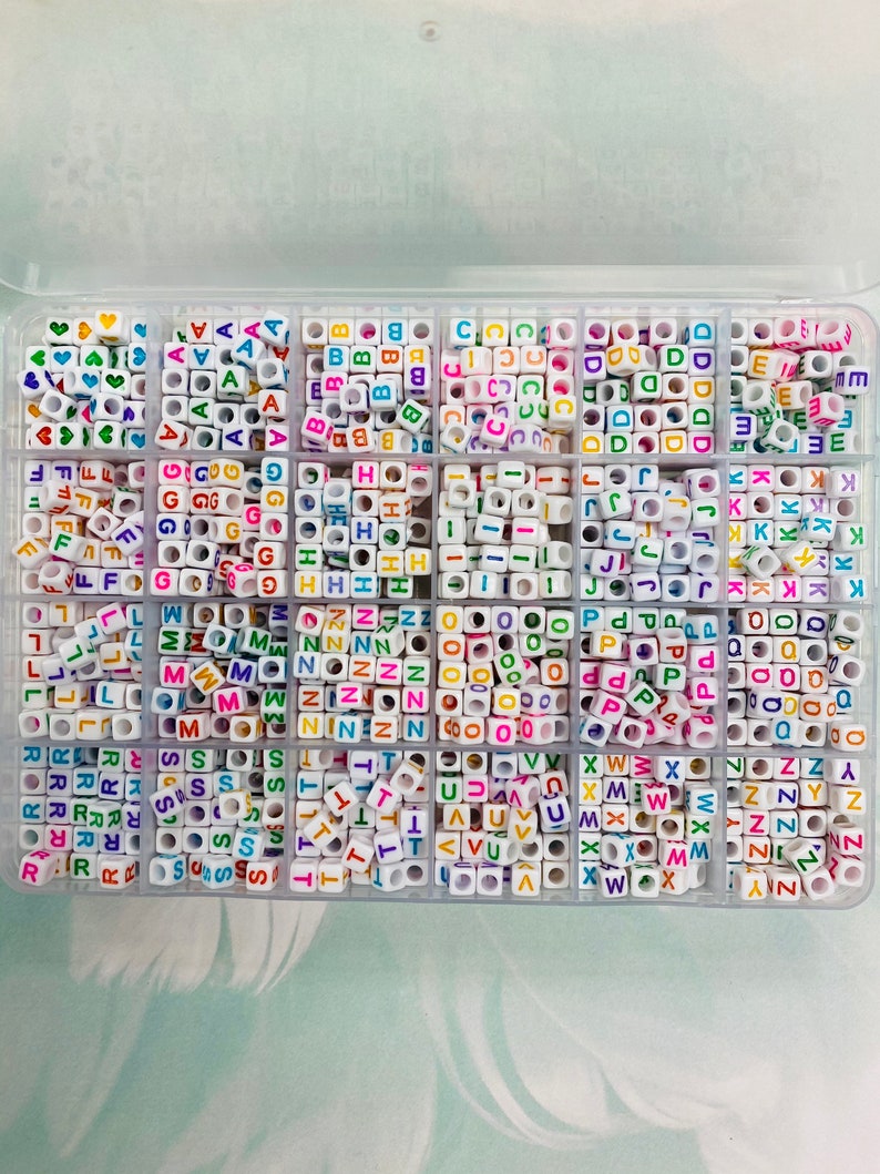 May include: A clear plastic storage box filled with small white letter beads. Each compartment contains a different letter of the alphabet, with the letters printed in various colors.