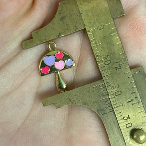 May include: A gold-colored mushroom charm with a loop for hanging. The mushroom cap is black with pink, red, and purple heart-shaped accents. The charm is being measured with a brass caliper.