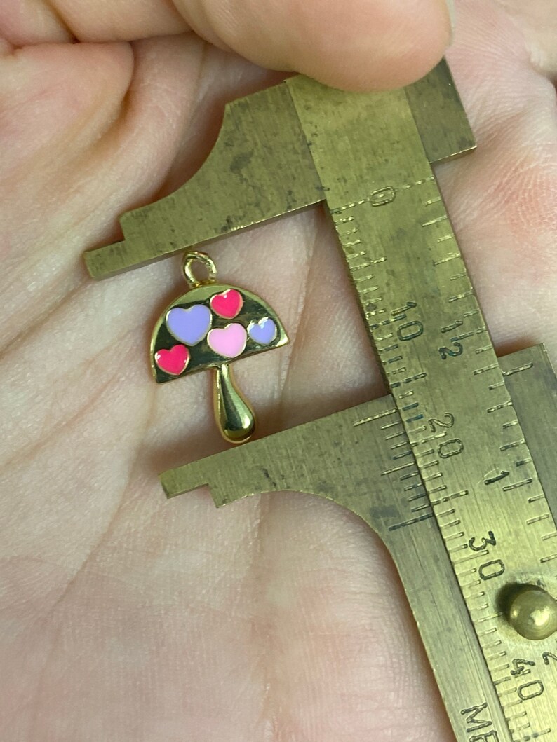 May include: A gold-colored mushroom charm with a loop for hanging. The mushroom cap is black with pink, red, and purple heart-shaped accents. The charm is being measured with a brass caliper.