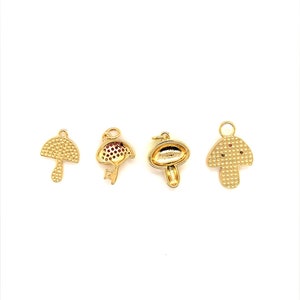 May include: Four gold-colored mushroom charms with varying designs. One charm has a textured cap and stem, while another features red accents. The charms are likely for jewelry making or crafting.
