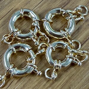 K&ouml;nnte beinhalten: Vier goldfarbene, runde Schmuckverschl&uuml;sse mit angebrachten Ketten. Jeder Verschluss hat ein kreisf&ouml;rmiges Design mit einem Scharnier und einer kleinen Schlaufe zur Befestigung. Die Verschl&uuml;sse sind auf einer Holzoberfl&auml;che angeordnet.