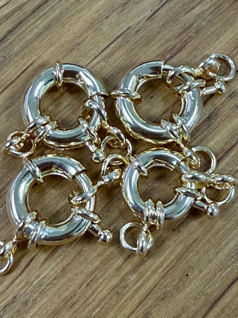 K&ouml;nnte beinhalten: Vier goldfarbene, runde Schmuckverschl&uuml;sse mit angebrachten Ketten. Jeder Verschluss hat ein kreisf&ouml;rmiges Design mit einem Scharnier und einer kleinen Schlaufe zur Befestigung. Die Verschl&uuml;sse sind auf einer Holzoberfl&auml;che angeordnet.