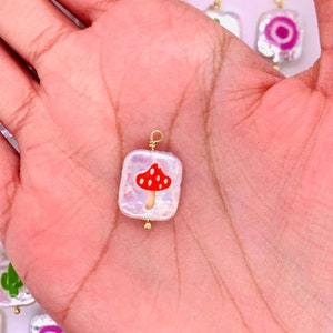 May include: A square, iridescent charm with a red mushroom design. The charm has a gold-colored wire loop and a small gold bead. Other charms with similar designs are visible in the background.