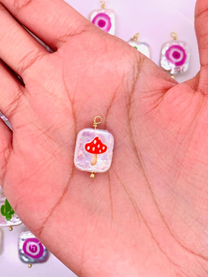 May include: A square, iridescent charm with a red mushroom design. The charm has a gold-colored wire loop and a small gold bead. Other charms with similar designs are visible in the background.