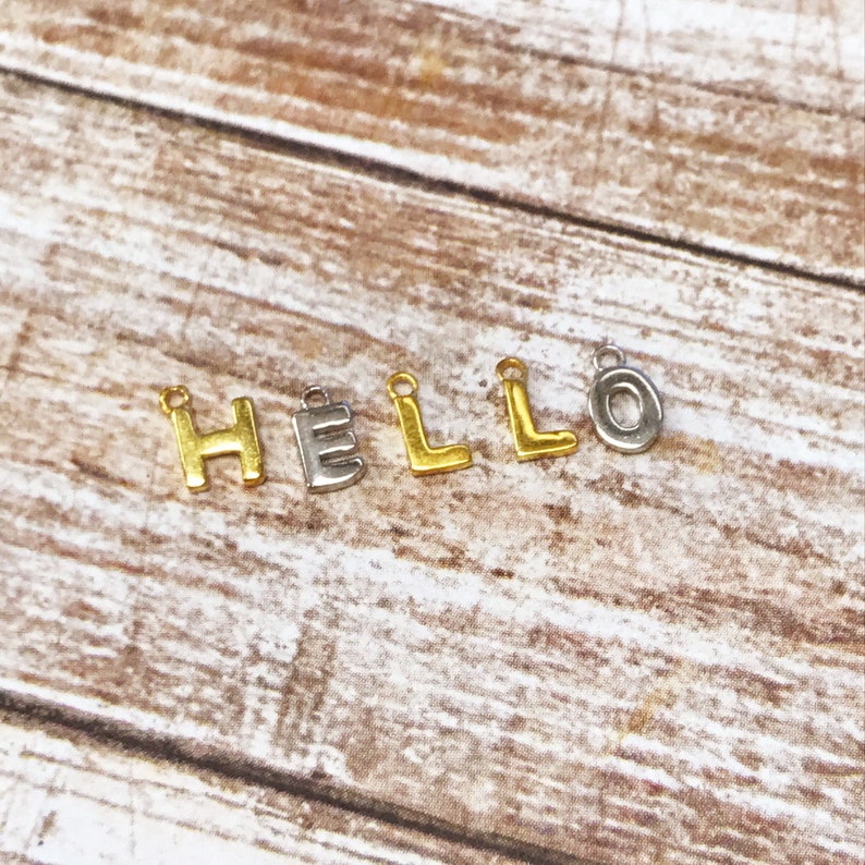 May include: Gold and silver letter charms spelling out the word "HELLO". The charms are on a wooden surface.