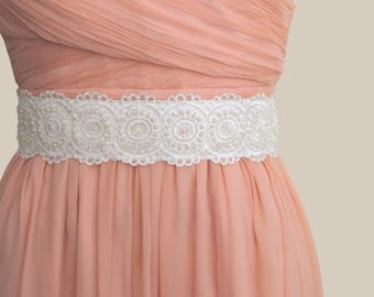 Könnte beinhalten: Ein pfirsichfarbenes Abendkleid mit einem weißen, perlenbesetzten Spitzenband. Der Gürtel hat ein sich wiederholendes kreisförmiges Muster mit perlenartigen Akzenten. Das Kleid hat ein drapiertes Oberteil und einen fließenden Rock.