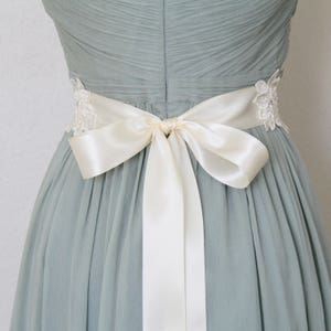 May include: A light blue dress with a cream-colored satin bow tied at the waist. The dress features delicate lace detailing on the sides and a pleated bodice, creating an elegant and feminine silhouette.