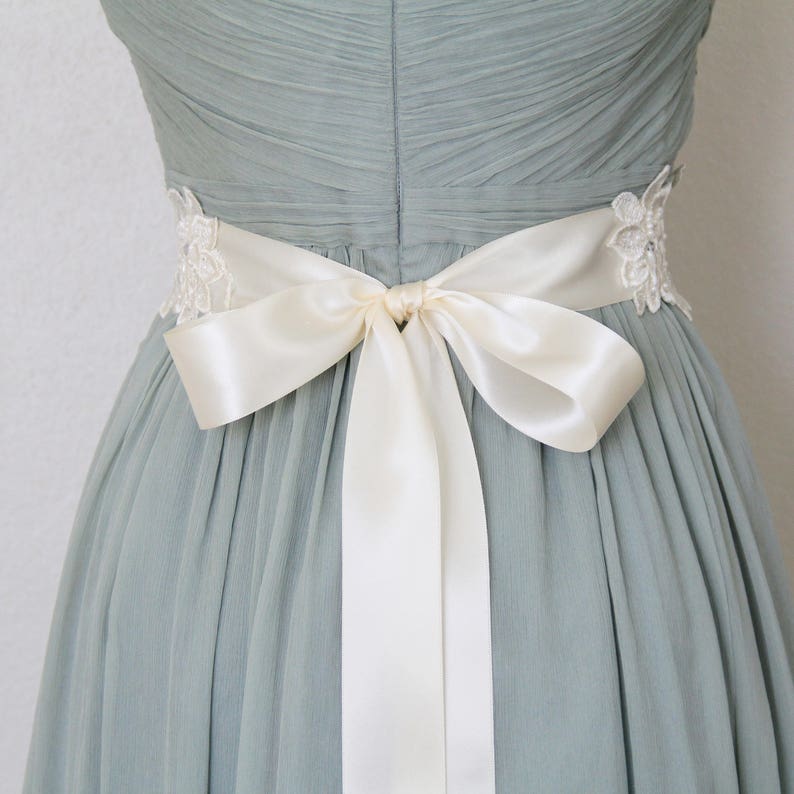 May include: A light blue dress with a cream-colored satin bow tied at the waist. The dress features delicate lace detailing on the sides and a pleated bodice, creating an elegant and feminine silhouette.