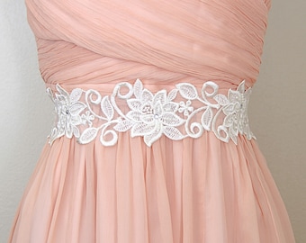 Könnte beinhalten: Ein hellpfirsichfarbenes formelles Kleid mit einem weißen floralen Spitzenband. Das Kleid hat ein plissiertes Oberteil und einen fließenden Rock. Der Gürtel ist mit zarten Blumen- und Blättermustern und kleinen Verzierungen verziert.