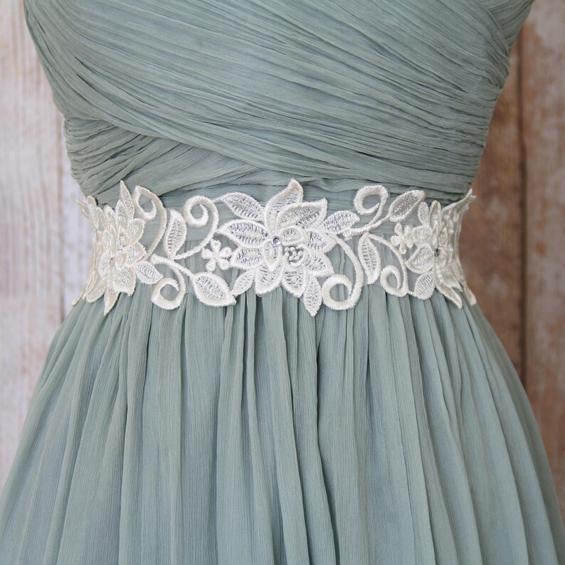 May include: A sage green dress with a white floral lace belt. The belt features a delicate design of flowers and leaves, with small embellishments. The dress has a pleated bodice and a flowing skirt, creating an elegant silhouette.