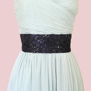 May include: A light blue dress with a dark navy blue embellished belt. The belt is decorated with intricate lace and sequins. The dress has a pleated skirt and a draped bodice. The background is a soft pink.