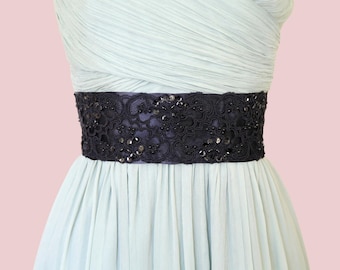 Könnte beinhalten: Ein hellblaues Kleid mit einem dunkelmarineblauen, verzierten Gürtel. Der Gürtel ist mit aufwendiger Spitze und Pailletten verziert. Das Kleid hat einen plissierten Rock und ein drapiertes Oberteil. Der Hintergrund ist ein zartes Rosa.