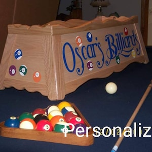 May include: A wooden pool table light fixture with the words "Oscar's Billiards" painted in blue. The fixture has a scalloped edge and is decorated with numbered billiard ball stickers. A set of billiard balls is in a wooden rack on a blue felt pool table.