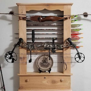 May include: A wooden wall-mounted bow rack with a drawer. The rack has a shelf for storing arrows and a hook for hanging a bow. There are two bows and a variety of arrows on the rack.