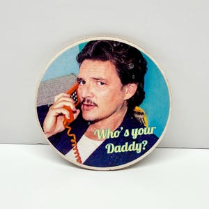 May include: Circular wooden coaster with a man on an orange telephone. The image has a retro style with a blue and teal background. The text on the coaster reads "Who's your Daddy?"