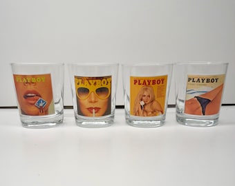 May include: A set of four clear glass tumblers, each featuring a different vintage Playboy magazine cover graphic. The designs include a close-up of lips, a face wearing yellow sunglasses, a blonde person, and a torso wearing black underwear.