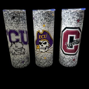 May include: Three glitter-covered tumblers with team logos. One features "ECU" in purple, another has the ECU pirate logo, and the third displays "gate" in maroon. Each tumbler has a silver glitter base and a blue lid.