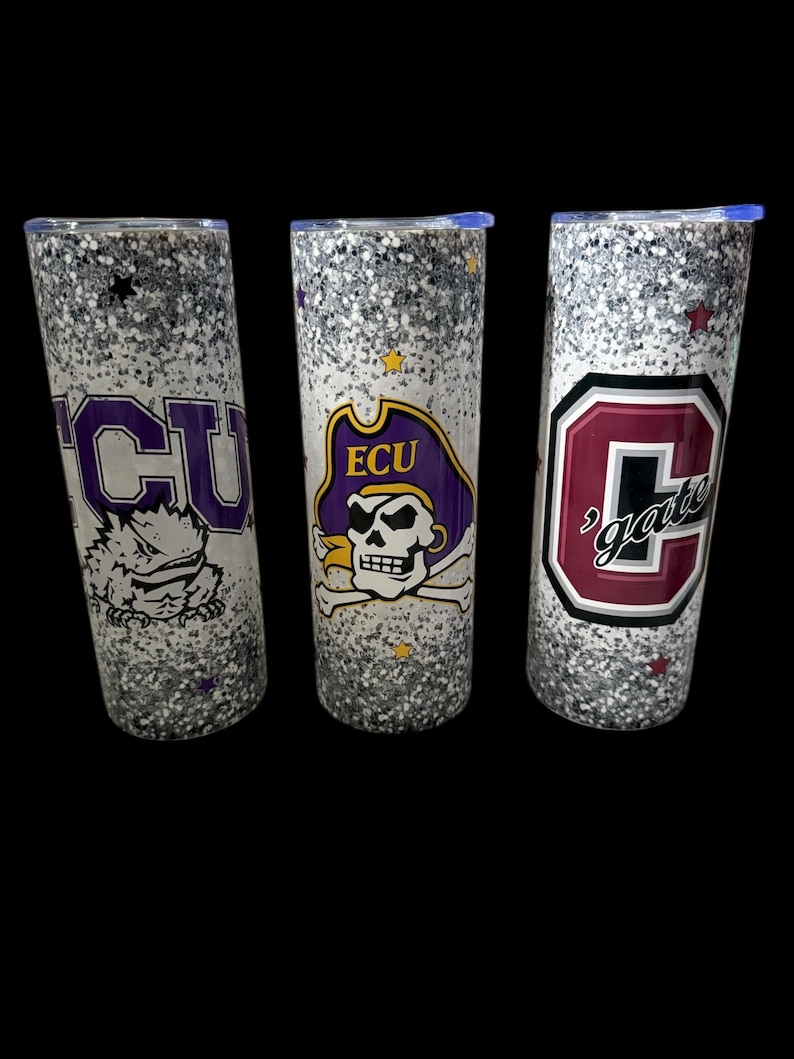 May include: Three glitter-covered tumblers with team logos. One features "ECU" in purple, another has the ECU pirate logo, and the third displays "gate" in maroon. Each tumbler has a silver glitter base and a blue lid.