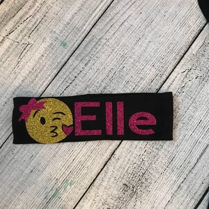 May include: Black headband with a pink glitter "Elle" and a yellow glitter emoji with a winking face and a pink heart.