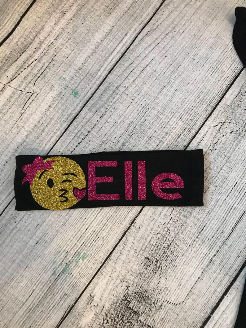May include: Black headband with a pink glitter "Elle" and a yellow glitter emoji with a winking face and a pink heart.