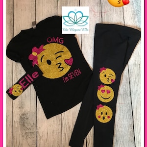 May include: A black t-shirt with a gold glitter emoji and the text "OMG I'm SEVEN". A matching black headband with a gold glitter emoji and the text "Elle". Black leggings with three gold glitter emojis.