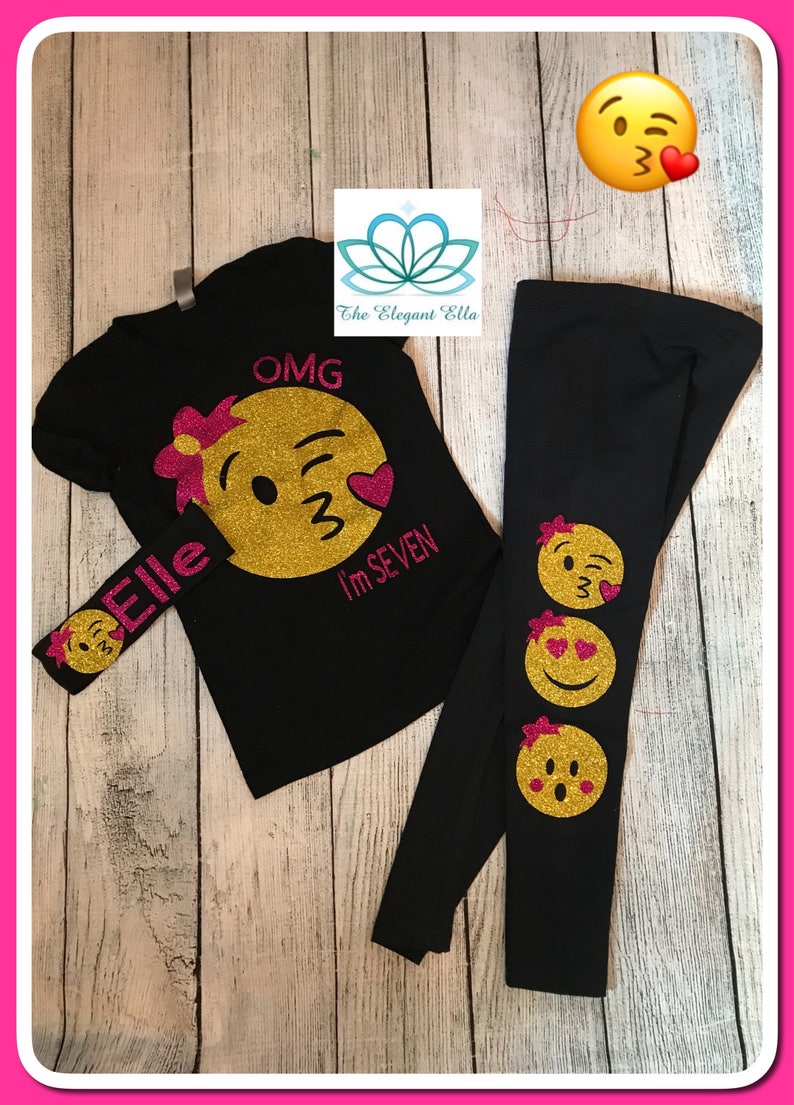 May include: A black t-shirt with a gold glitter emoji and the text "OMG I'm SEVEN". A matching black headband with a gold glitter emoji and the text "Elle". Black leggings with three gold glitter emojis.