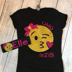 May include: Black t-shirt with a gold glitter winking emoji with a pink bow and the text "OMG I'm SEVEN". A matching headband with a winking emoji and the text "Elle" is included.