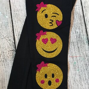 May include: Black leggings with three gold glitter emojis with pink bows and pink hearts. The emojis are a winking kiss, heart eyes, and a surprised face.