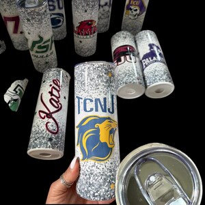 May include: A collection of glitter-covered, insulated tumblers. Each tumbler features a different college or university logo and name, including "TCNJ", "ECU", and "Katie". The tumblers are cylindrical and have a silver and white glitter design.