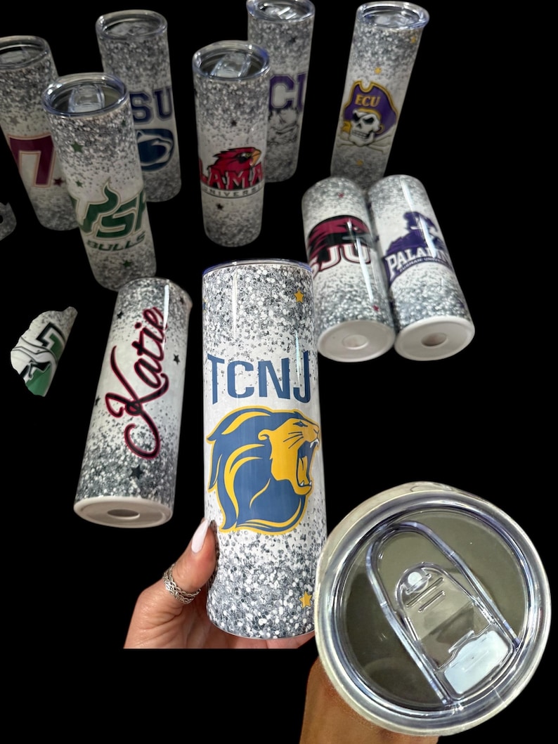 May include: A collection of glitter-covered, insulated tumblers. Each tumbler features a different college or university logo and name, including "TCNJ", "ECU", and "Katie". The tumblers are cylindrical and have a silver and white glitter design.