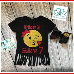 May include: Black t-shirt with a gold glitter winking emoji with a red bow and red heart. The text "Birthday Girl" and "Giuliana 7" are in red glitter. The shirt has a frayed bottom.