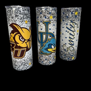 May include: Three glitter-covered tumblers with black lids. One features a brown and yellow owl logo with the letters "RU". Another has a blue and yellow logo with the letters "UP" and a cartoon roadrunner. The third has the name "Emily" in blue script.