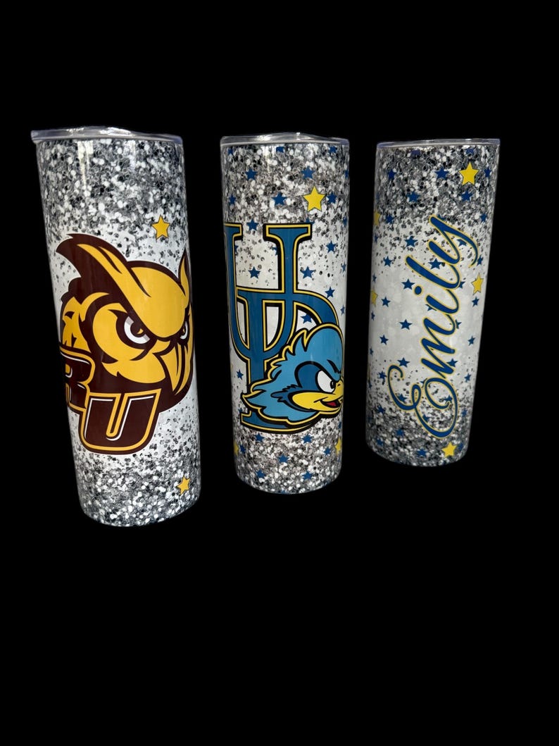May include: Three glitter-covered tumblers with black lids. One features a brown and yellow owl logo with the letters "RU". Another has a blue and yellow logo with the letters "UP" and a cartoon roadrunner. The third has the name "Emily" in blue script.