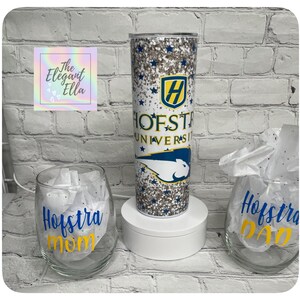 May include: A glitter-covered tumbler with the Hofstra University logo and text, alongside two wine glasses with "Hofstra Mom" and "Hofstra Dad" in blue and yellow. The tumbler is cylindrical and tall, while the glasses are stemless.