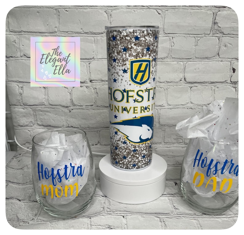 May include: A glitter-covered tumbler with the Hofstra University logo and text, alongside two wine glasses with "Hofstra Mom" and "Hofstra Dad" in blue and yellow. The tumbler is cylindrical and tall, while the glasses are stemless.