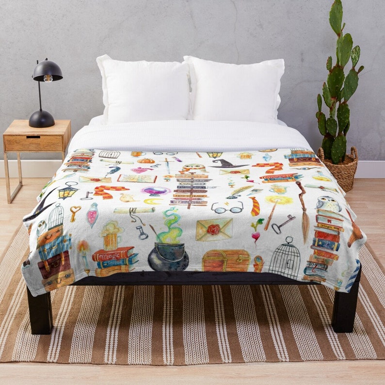 May include: A white throw blanket with a watercolor pattern of various magical objects, including books, owls, wands, potions, and a cauldron. The pattern is on a white background and is surrounded by a white border.
