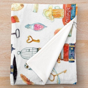 May include: White fleece throw blanket with a watercolor pattern of various objects including a beer mug, a birdcage, a key, a feather, a potion bottle, a hat, and books with the words "Magic" and "Poison" on the spines.