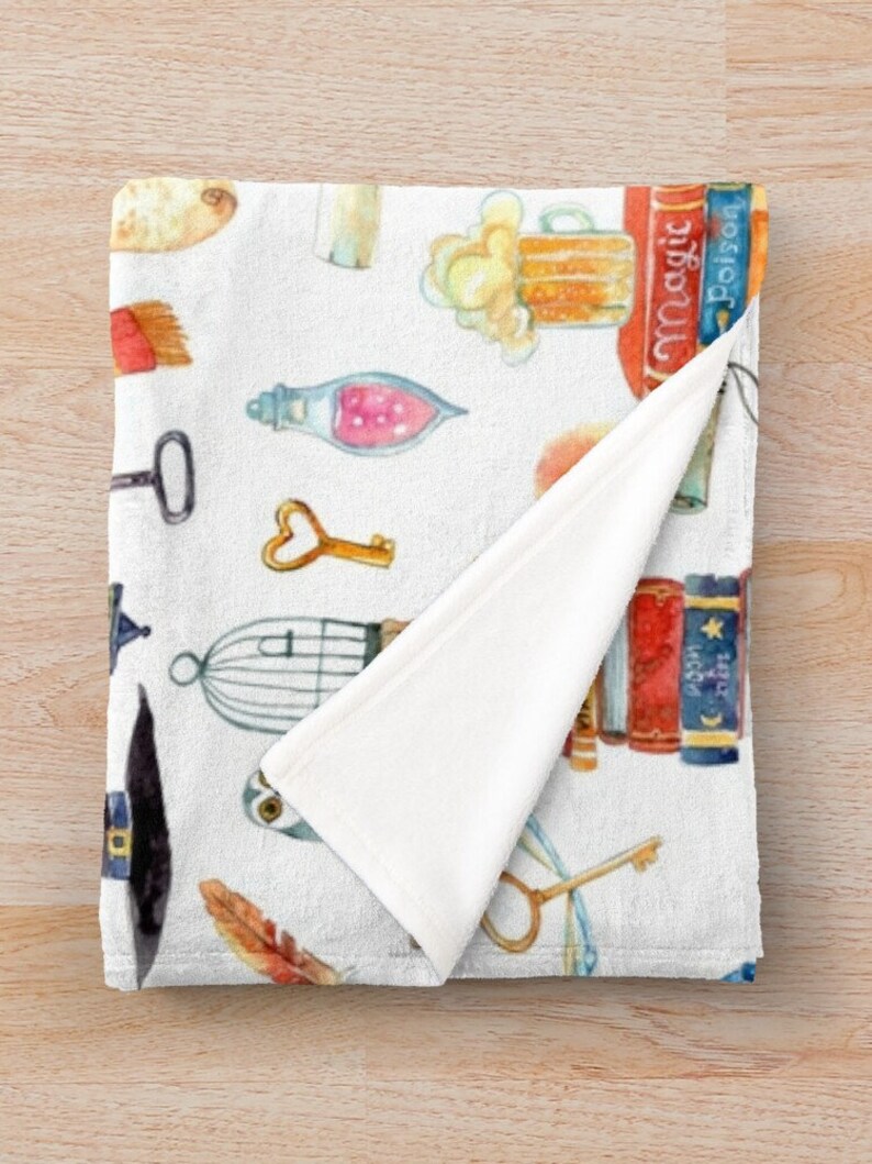 May include: White fleece throw blanket with a watercolor pattern of various objects including a beer mug, a birdcage, a key, a feather, a potion bottle, a hat, and books with the words "Magic" and "Poison" on the spines.