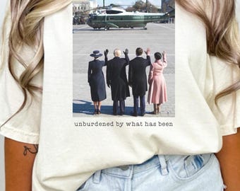 Peut inclure: Un t-shirt blanc avec une photo de quatre personnes faisant signe au revoir alors qu'un hélicoptère survole. Le texte "unburdened by what has been" est imprimé sous la photo.